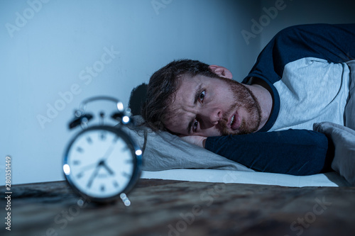 Desperate stressed young man whit insomnia lying in bed staring at alarm clock trying to sleep photo
