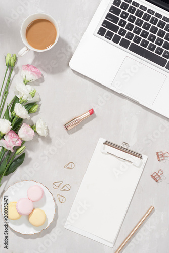 Feminine flat lay workspace with laptop, cup of tea, macarons, lipstick and flowers on white table. Top view mock up.