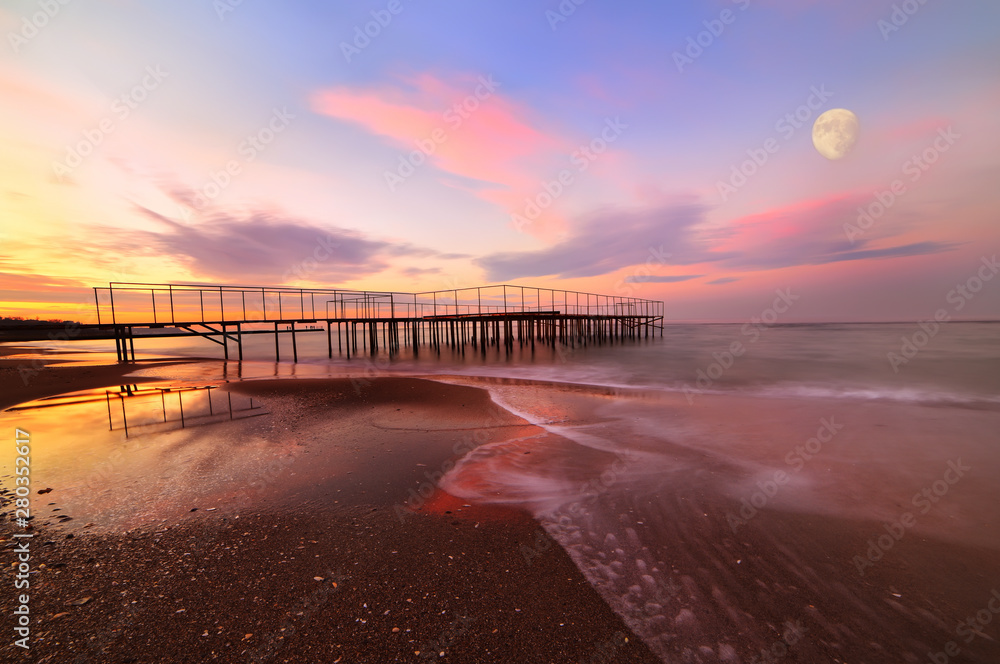 Deserted beach at sunset. a huge moon above the water and an old iron pier.