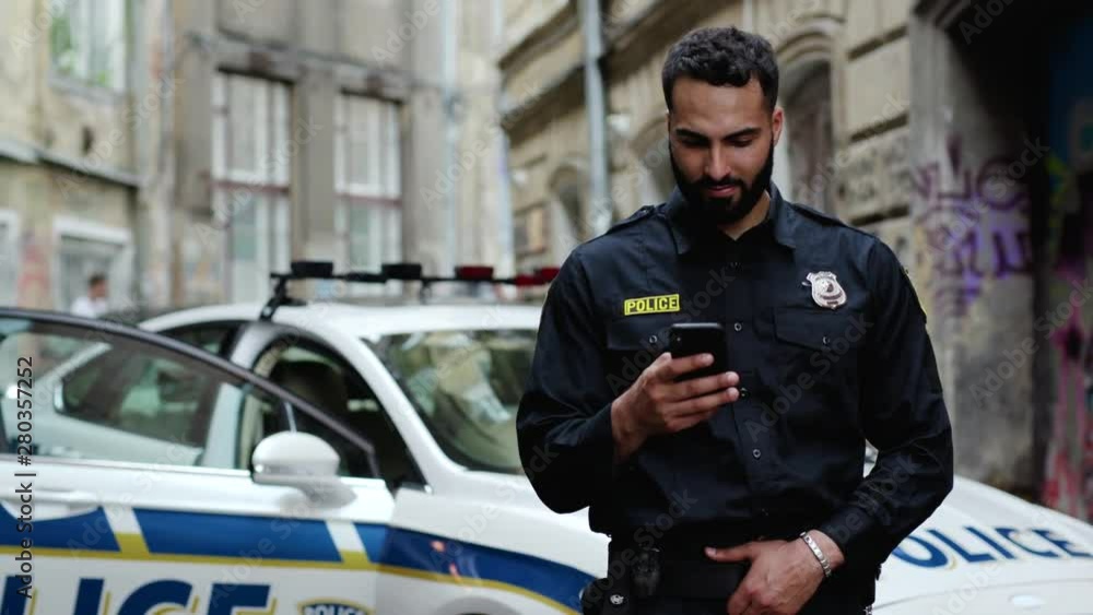 Attractive police officer on duty using a mobile phone typing message to friend standing outdoors on background of police patrol car in the street. Attractive police officer on duty using a mobile phone typing message to friend standing outdoors on background of police patrol car in the street.