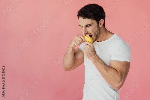 hungry muscular man in white t-shirt eating banana isolated on pink