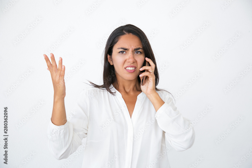 Annoyed beautiful woman speaking on phone. Angry young Latin woman in white shirt making displeased face and gestures and talking on cell. Feeling stressed concept Annoyed beautiful woman speaking on phone. Angry young Latin woman in white shirt making displeased face and gestures and talking on cell. Feeling stressed concept