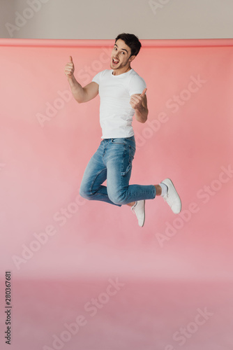 Fototapeta Naklejka Na Ścianę i Meble -  smiling muscular man in white t-shirt and jeans jumping and showing thumbs up on pink