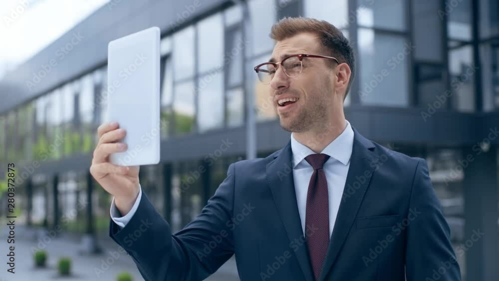 businessman with digital tablet having video call, showing peace sign, talking, smiling and gesturing near building businessman with digital tablet having video call, showing peace sign, talking, smiling and gesturing near building
