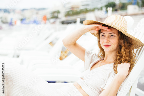 Beautiful ,elegant, teenager, with red hair and freckles,wearing a white dress and a hat, enjoy the seaside resort, watch in the distance the beautiful panorama covering her eyes with the palm of hand © jon11