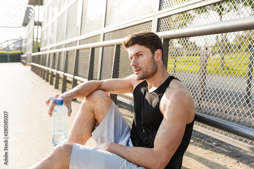 Portrait of attractive man drinking water while sitting on sports ground during morning workout outdoors