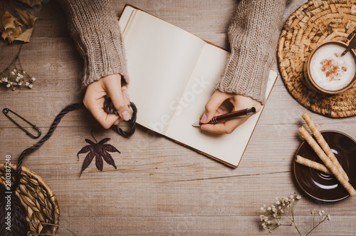 Fall winter background. Female hand holding a pen for a sketch or letter on old notebook with a ball of wool yarn and cup of cappuccino on a wooden background. Home hobbies, Cozy comfort concept. photo