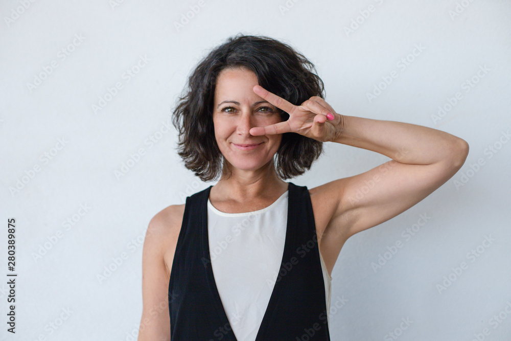Happy woman showing peace sign. Beautiful middle aged woman with curly hair showing victory sign and smiling at camera isolated on grey background. Gesture concept Happy woman showing peace sign. Beautiful middle aged woman with curly hair showing victory sign and smiling at camera isolated on grey background. Gesture concept