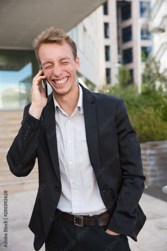Joyful manager chatting on phone outside during work break. Happy young man in office suit standing near city building, talking on cell and laughing. Communication and fun concept Joyful manager chatting on phone outside during work break. Happy young man in office suit standing near city building, talking on cell and laughing. Communication and fun concept
