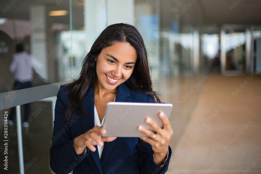 Fototapeta premium Cheerful happy office worker using tablet in office hall. Young Latin business woman leaning on glass wall and reading on screen. Tablet computer concept