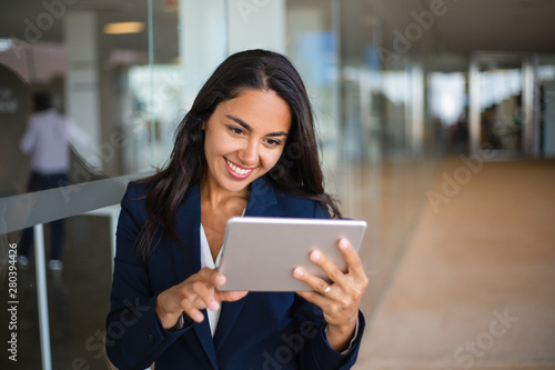 Cheerful happy office worker using tablet in office hall. Young Latin business woman leaning on glass wall and reading on screen. Tablet computer concept