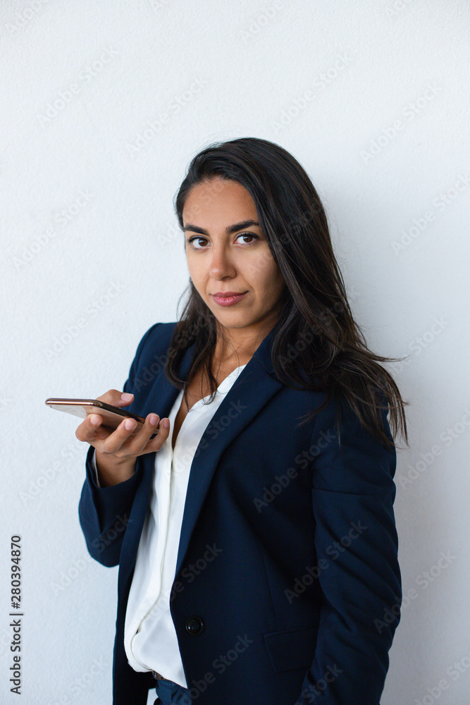 Confident businesswoman holding mobile phone. Attractive young brunette woman in formal wear using smartphone and looking at camera. Technology concept Confident businesswoman holding mobile phone. Attractive young brunette woman in formal wear using smartphone and looking at camera. Technology concept