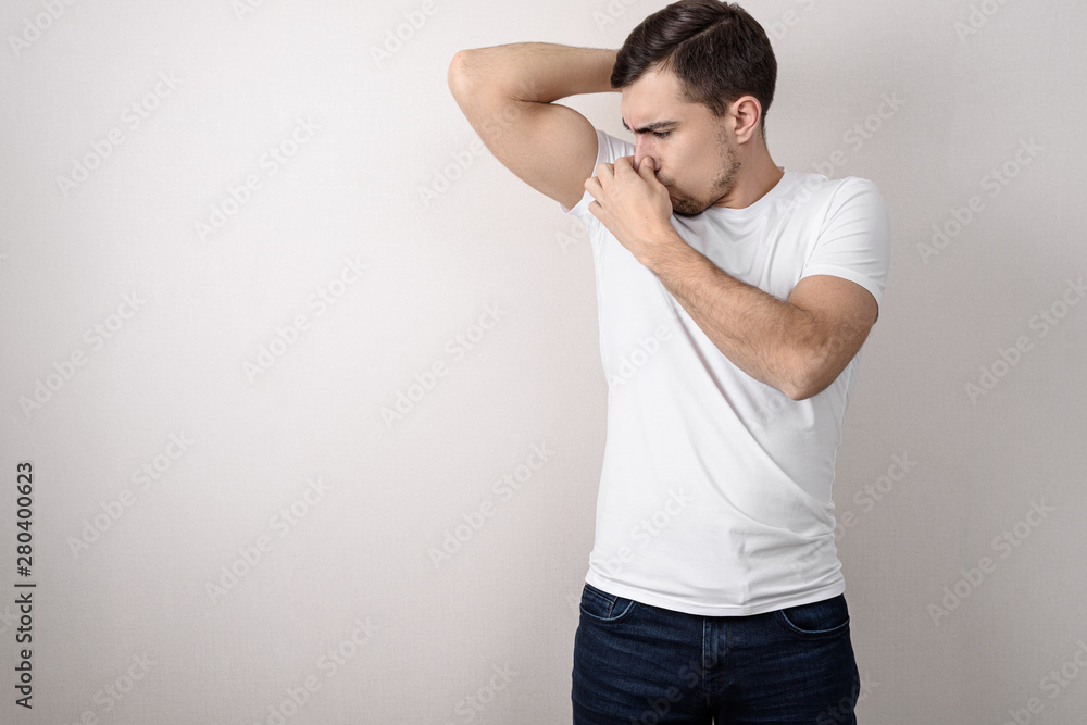young guy in white t-shirt clamps his nose from smell of sweat from his armpits on gray background with copy space young guy in white t-shirt clamps his nose from smell of sweat from his armpits on gray background with copy space