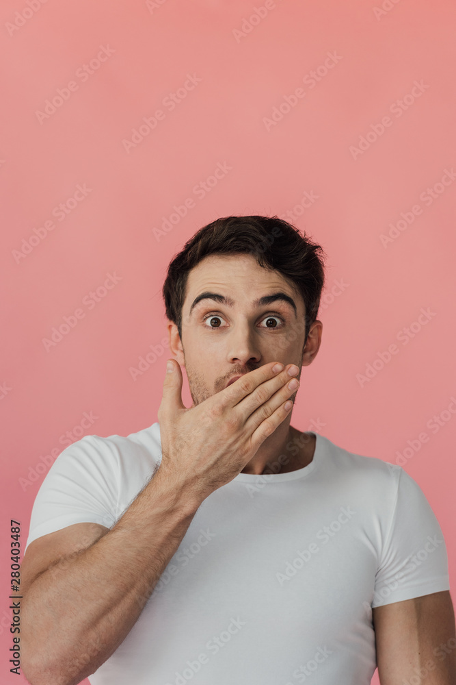 front view of shocked muscular man in white t-shirt covering mouth with hand isolated on pink front view of shocked muscular man in white t-shirt covering mouth with hand isolated on pink