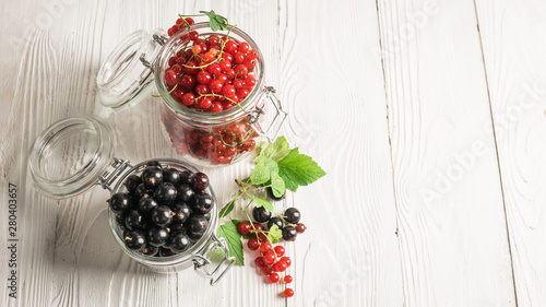 Fototapeta Naklejka Na Ścianę i Meble -  Ripe fresh black and red currant berries in glass jars on white wooden background. Horizontal frame. Selective focus.