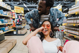 selective focus of happy asian girl sunglasses and holding bottle with wine while sitting in shopping cart near cheerful african american man in store