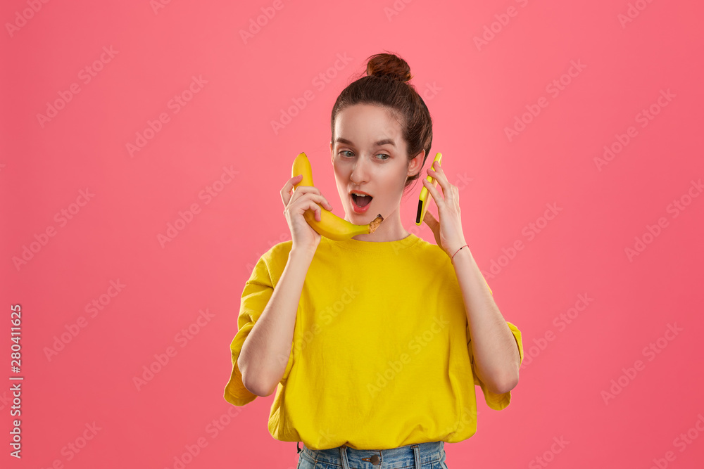 Smiling model with a hair bun in a yellow t-shirt holding banana speaking on a yellow smartphone. Smiling model with a hair bun in a yellow t-shirt holding banana speaking on a yellow smartphone.