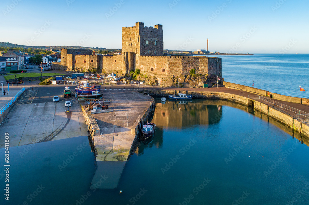 Obraz premium Medieval Norman Castle and harbor with boat ramp and wave breaker in Carrickfergus near Belfast, Northern Ireland, UK. Aerial view in sunset light. Old power plant in the background