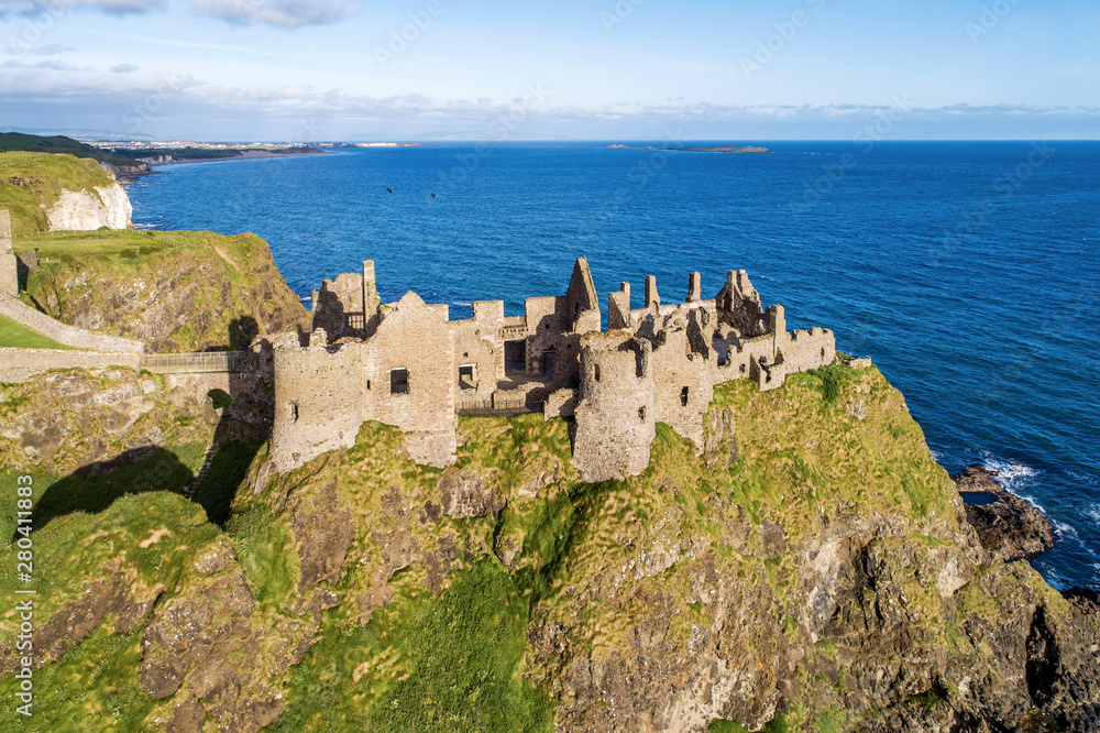 Ruins of medieval Dunluce Castle on a steep cliff near Bushmills. Northern coast of County Antrim, Northern Ireland, UK. Aerial view in sunrise light. Far view of Portrush resort in the background