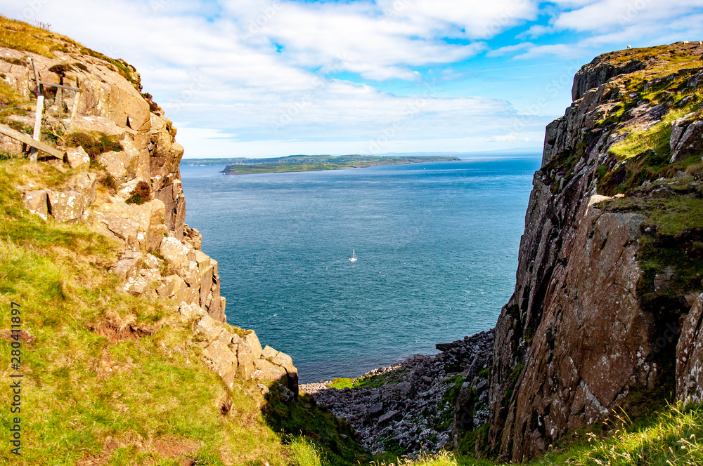 Northern Ireland, UK. Fragment of Fair Head cliff at the north-eastern corner of County Antrim with the far view of Rathlin Island, a sailboat and Atlantic Ocean