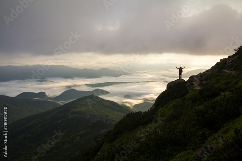 Wide mountain panorama. Small silhouette of tourist with backpack on rocky mountain slope with raised hands over valley covered with white puffy clouds. Beauty of nature, tourism and traveling concept