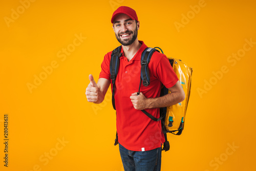Photo of unshaven delivery man in red uniform showing thumb up and carrying backpack with takeaway food isolated over yellow background
