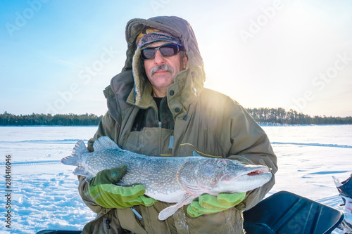 male fisherman with a mustache in sunglasses is holding a big fish pike in the winter