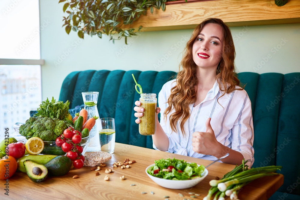 Plakat Young woman drinking smoothie in the beautiful interior with green flowers on the background and fresh fruits and vegetables on the table. Healthy eating concept. Vegan meal and detox menu