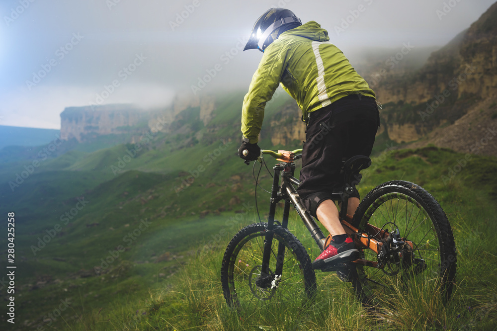 Fototapeta premium Aged male athlete in helmet and mask rides a mountain bike on a grassy slope against the background of plateau rocks and low clouds on an overcast day. Downhill mountain bike concept
