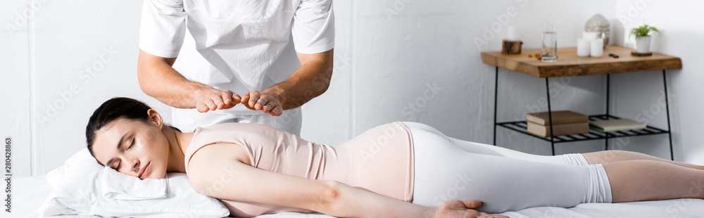 Fototapeta premium panoramic shot of healer standing near woman lying with closed eyes on massage table and holding hands above her body