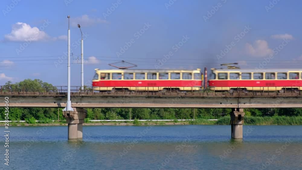 Tram moves along the bridge over the river on a background of industrial plant and blue cloudy sky. Tram moves along the bridge over the river on a background of industrial plant and blue cloudy sky.