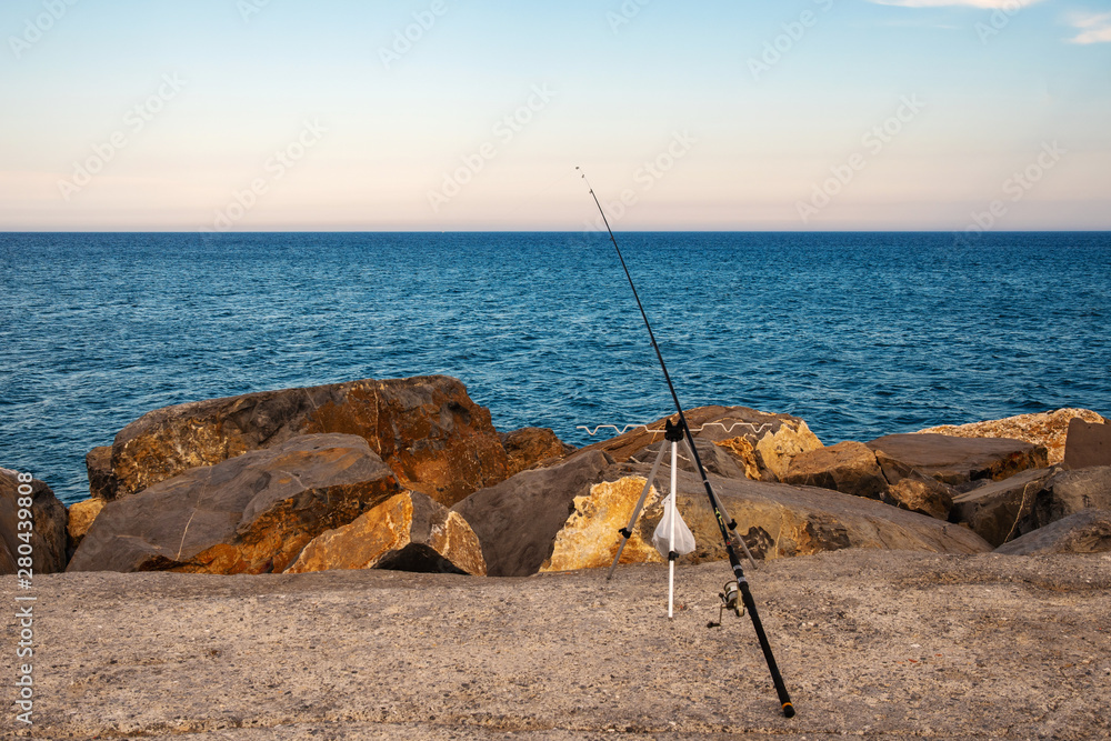 A fishing rod placed on the rocky quayside on the shore of the sea at sunset, Diano Marina, Imperia, Liguria, Italy A fishing rod placed on the rocky quayside on the shore of the sea at sunset, Diano Marina, Imperia, Liguria, Italy
