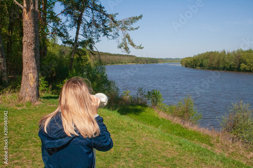 Young woman looking through binoculars, forest, river and roofs of the city on background photo
