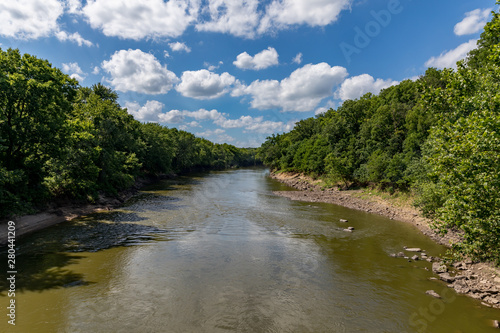 Water flowing along rocky riverbank of Sangamon River surrounded by trees on a sunny day with clouds