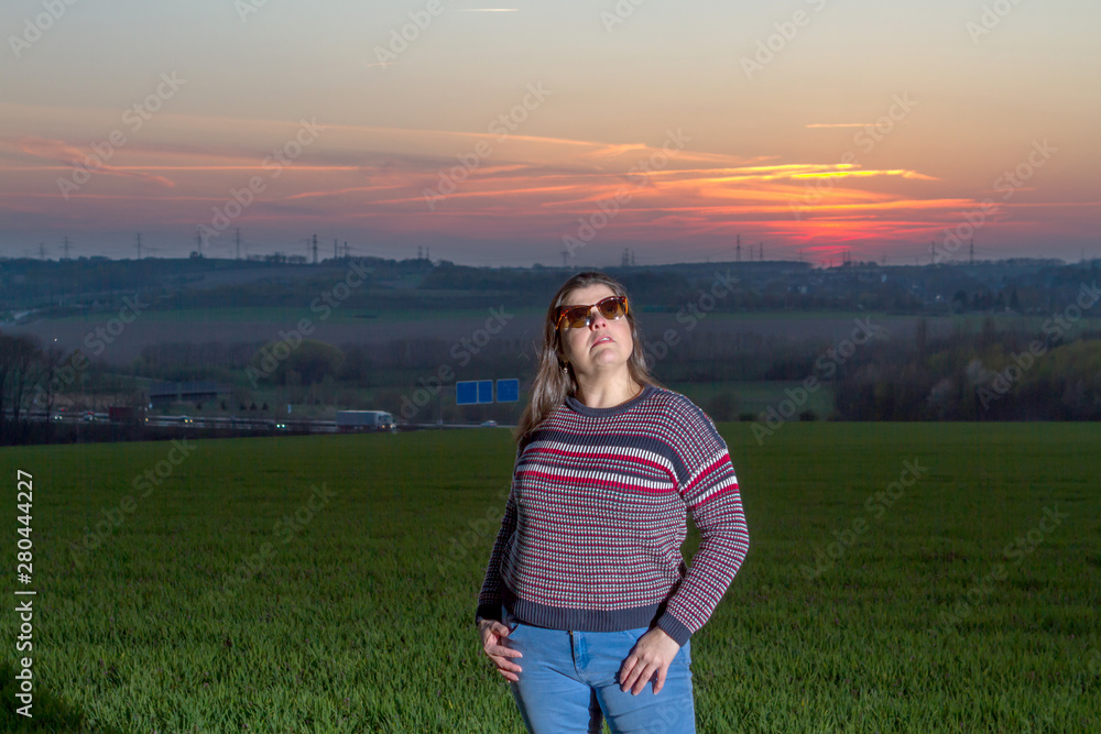 Lady poses outside in the field during twilight. She is wearing sunglasses and is looking away.