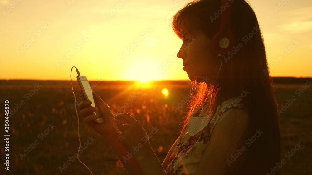 girl listening to music and dancing in the rays of a beautiful sunset. young girl in headphones and with a smartphone touches finger to the sensor of the tablet selects songs online. Slow motion.