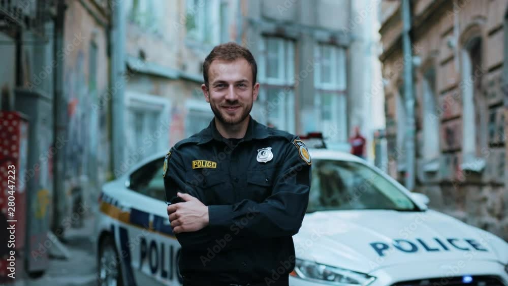 Smiling young man cops stand near patrol car look at camera wear glasses enforcement happy officer police uniform auto safety security communication control policeman close up slow motion Smiling young man cops stand near patrol car look at camera wear glasses enforcement happy officer police uniform auto safety security communication control policeman close up slow motion