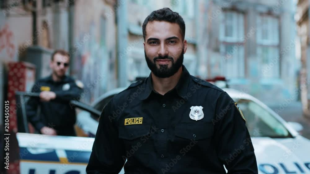 Strong smiling young man cops stand near patrol car look at camera enforcement officer police uniform auto safety security communication control policeman close up slow motion Strong smiling young man cops stand near patrol car look at camera enforcement officer police uniform auto safety security communication control policeman close up slow motion