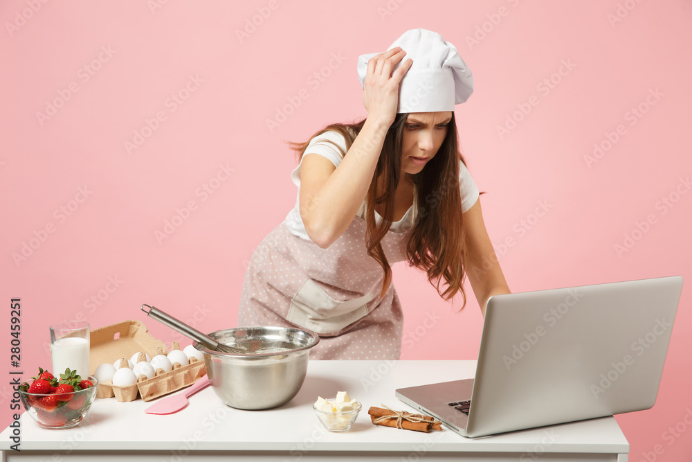 Chef cook confectioner or baker in apron white t-shirt, toque chefs hat cooking Christmas cake or cupcake at table looking for recipe on pc laptop isolated on pink pastel background. Mock up concept. Chef cook confectioner or baker in apron white t-shirt, toque chefs hat cooking Christmas cake or cupcake at table looking for recipe on pc laptop isolated on pink pastel background. Mock up concept.
