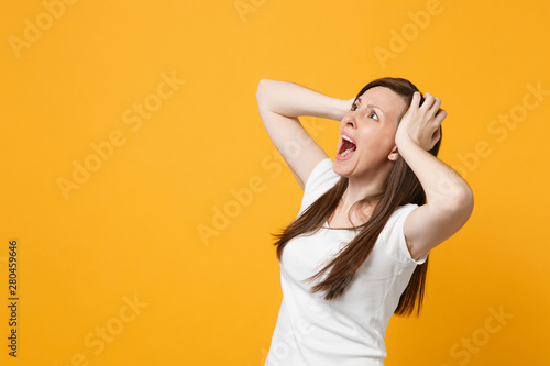 Portrait of screaming young woman in white casual clothes looking aside, putting hands on head isolated on bright yellow orange wall background in studio. People lifestyle concept. Mock up copy space.