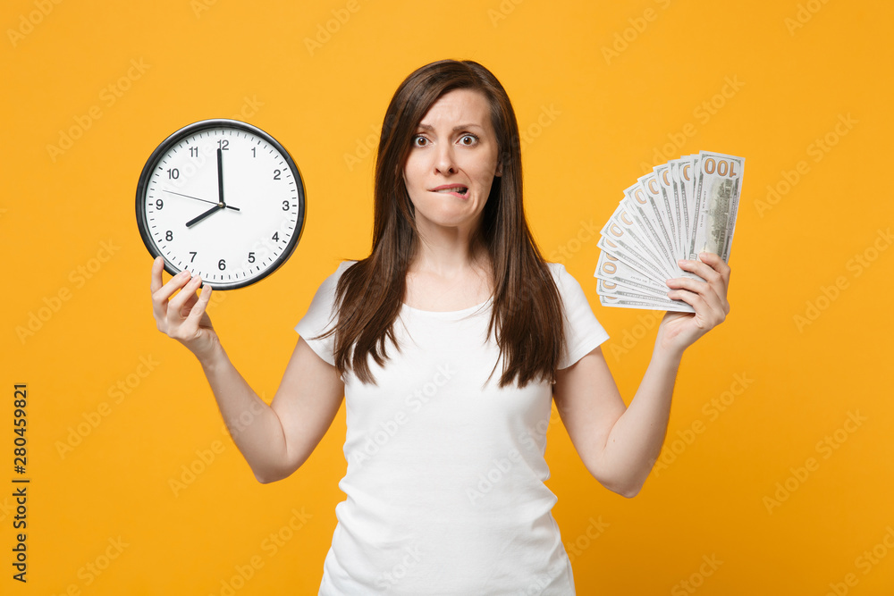 Nervous young woman in white casual clothes biting lips, holding round  clock, fan of cash money in dollar banknotes isolated on yellow orange  background. People lifestyle concept. Mock up copy space. Stock