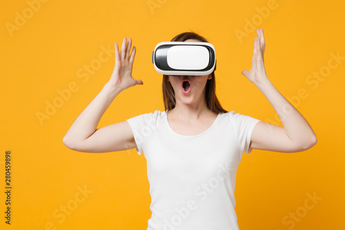 Shocked young woman in white casual clothes watching in headset of virtual reality, spreading hands isolated on bright yellow orange background in studio. People lifestyle concept. Mock up copy space.