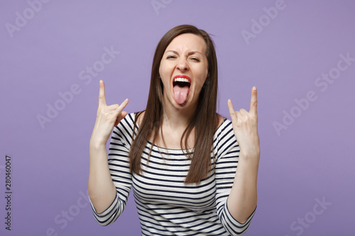 Crazy young woman in casual striped clothes posing isolated on violet purple background. People lifestyle concept. Mock up copy space. Depicting heavy metal rock sign horns up gesture, showing tongue.