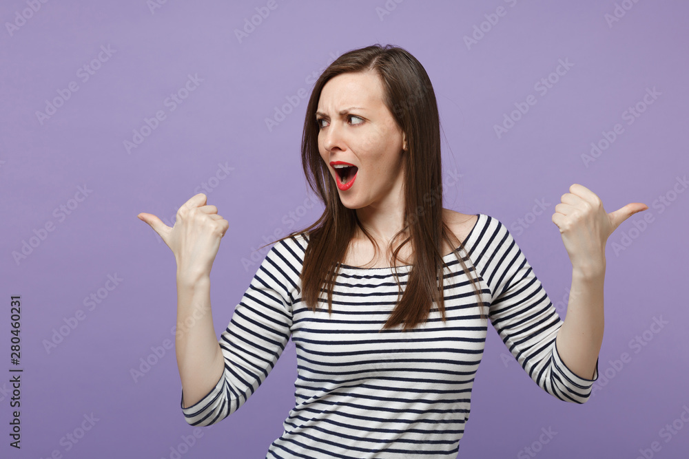 Excited perplexed young brunette woman girl in casual striped clothes posing isolated on violet purple background studio portrait. People lifestyle concept. Mock up copy space. Pointing thumbs aside. Excited perplexed young brunette woman girl in casual striped clothes posing isolated on violet purple background studio portrait. People lifestyle concept. Mock up copy space. Pointing thumbs aside.