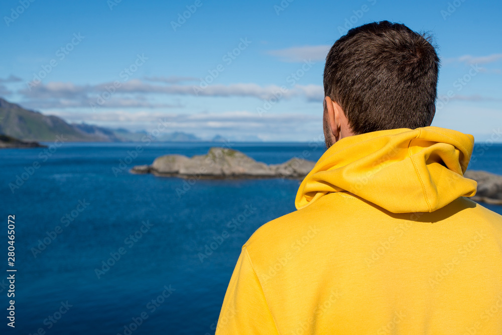 Fototapeta premium A young man in a yellow hoodie outdoor enjoy the northern beauty. Ocean and rocks landscape. Scenic view. Travel, adventure. Sense of freedom, lifestyle. Lofoten Islands, Norway. Summer in Scandinavia