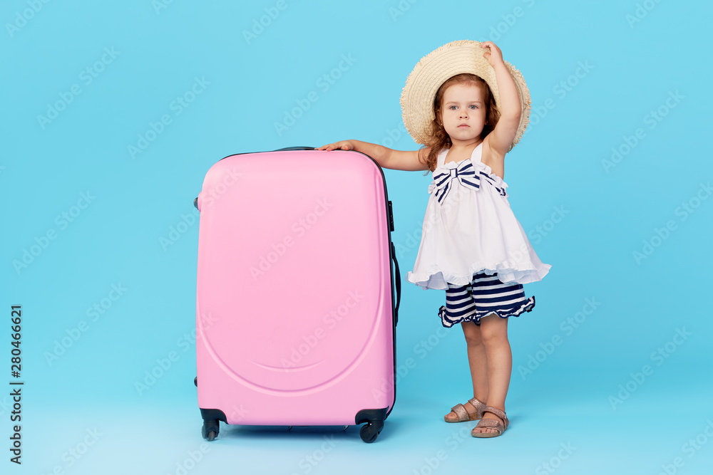 © oes - Happy child girl tourist packs clothes into a suitcase for travel, summer vacation. Toddler kid in straw hat  holding a pink suitcase on a blue background