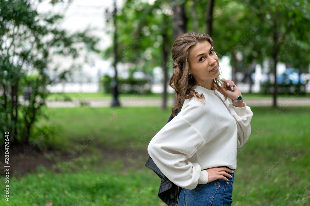 Happy Caucasian girl. Young beautiful European long free hair walks in city park in cheerful mood. Smile, white teeth. Wearing pullover. In hands black leather jacket. To enjoy life. Lifestyle Happy Caucasian girl. Young beautiful European long free hair walks in city park in cheerful mood. Smile, white teeth. Wearing pullover. In hands black leather jacket. To enjoy life. Lifestyle