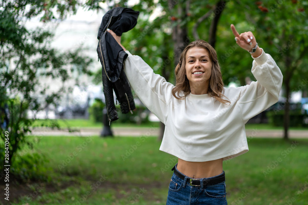 Lucky girl. Young beautiful Caucasian girl long curly hair walks in city park in great mood. Smile, white teeth. Wearing pullover. In hands black leather jacket. enjoy life. Lifestyle Lucky girl. Young beautiful Caucasian girl long curly hair walks in city park in great mood. Smile, white teeth. Wearing pullover. In hands black leather jacket. enjoy life. Lifestyle