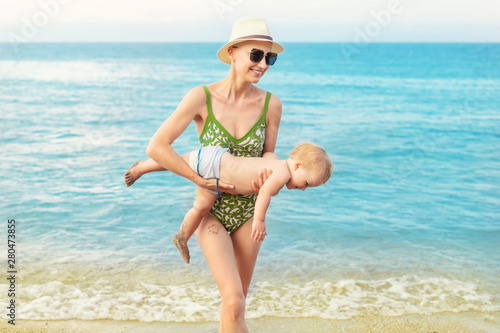 Canvas Print Young beautiful bald mother carrying cute caucasian toddler boy out clear blue water at tropical beach with warm summer rain