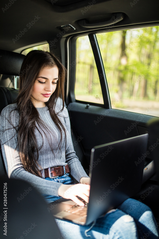 Beautiful s woman is using a laptop and smiling while sitting on back seat in the car